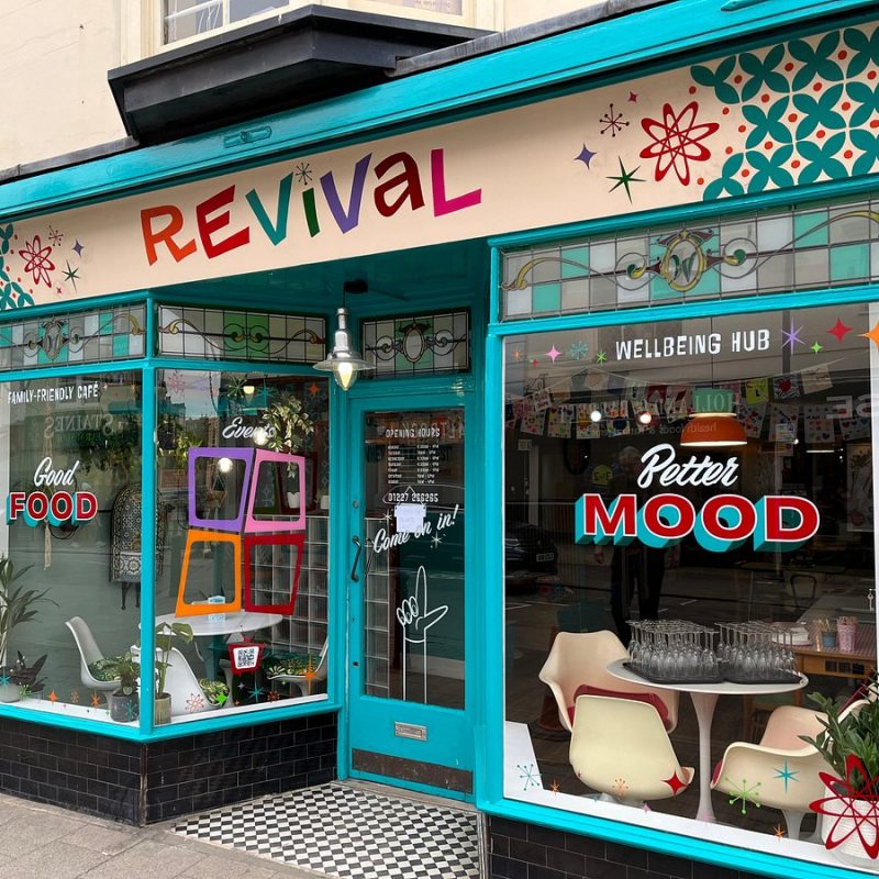 Front view of the Revival café and wellbeing hub in Whitstable, with a bright turquoise shopfront and large windows decorated with colourful graphics. The signs read “Good Food”, “Better Mood” and “Wellbeing Hub”, with chairs and tables visible inside.