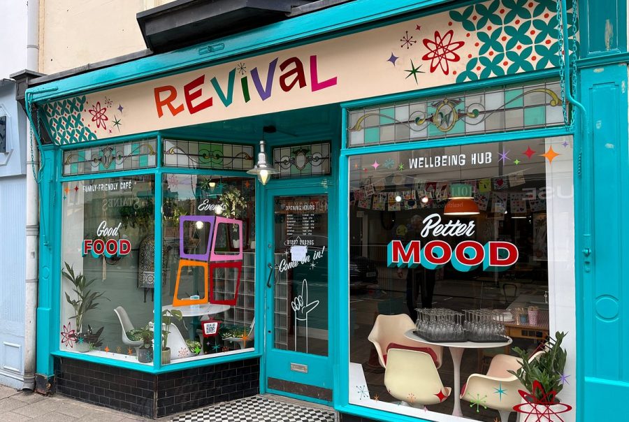 Front view of the Revival café and wellbeing hub in Whitstable, with a bright turquoise shopfront and large windows decorated with colourful graphics. The signs read “Good Food”, “Better Mood” and “Wellbeing Hub”, with chairs and tables visible inside.