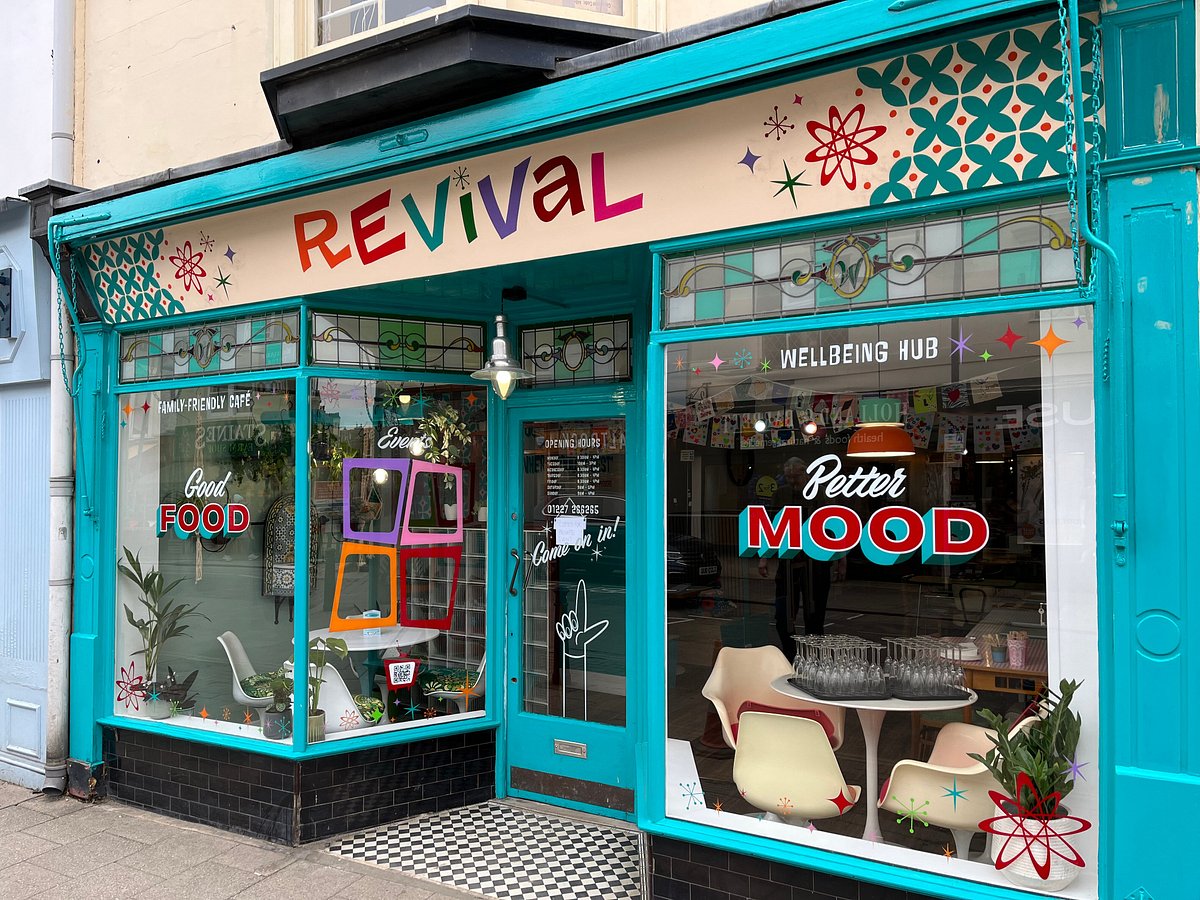 Front view of the Revival café and wellbeing hub in Whitstable, with a bright turquoise shopfront and large windows decorated with colourful graphics. The signs read “Good Food”, “Better Mood” and “Wellbeing Hub”, with chairs and tables visible inside.